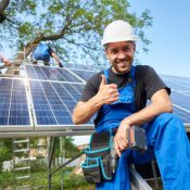 Portrait of smiling technician with electrical screwdriver showing thumb-up in front of unfinished high exterior solar panel photo voltaic system with team of workers on high platform.